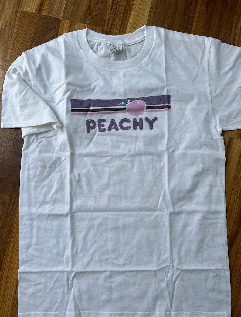 May include: White t-shirt with a graphic design featuring a peach illustration and the word "PEACHY" in purple letters. The design includes a horizontal stripe in shades of purple and brown.