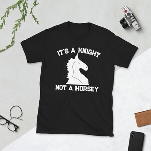 Puede incluir: Camiseta negra con un diseño de caballo de ajedrez blanco y el texto "IT'S A KNIGHT NOT A HORSEY". Una cámara, una cartera, un teléfono y gafas son visibles en el fondo.