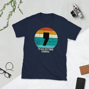 May include: A navy blue t-shirt with a retro design. The design features a black comma inside a circle with orange, yellow, teal, and white stripes. The text "TEAM OXFORD COMMA" is below the circle.