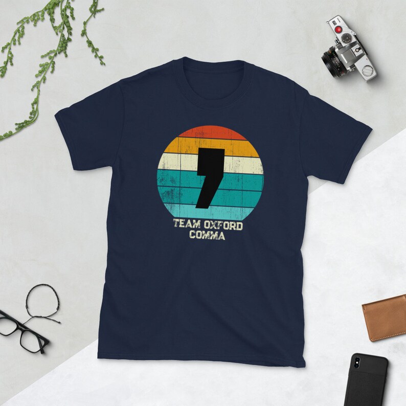May include: A navy blue t-shirt with a retro design. The design features a black comma inside a circle with orange, yellow, teal, and white stripes. The text "TEAM OXFORD COMMA" is below the circle.