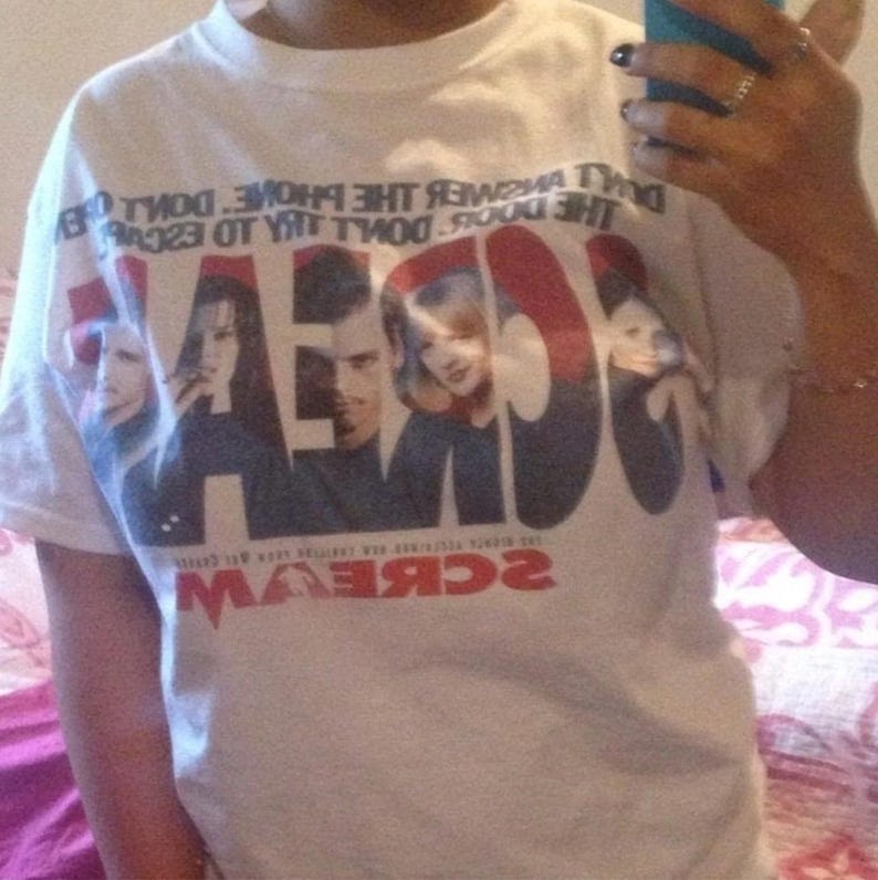 May include: White t-shirt featuring the word "SCREAM" in large, bold letters with images of actors from the movie. The shirt also has text around the top of the design. The design is in red, black, and white.