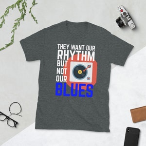 May include: A dark heather grey t-shirt with a white graphic print that says "They want our rhythm but not our blues". The graphic also includes a red circle with a black record player inside.