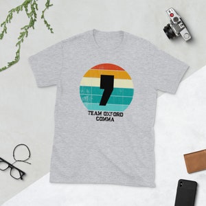May include: A light grey t-shirt with a retro design. It features a black comma inside a circle of orange, yellow, teal, and white stripes. The text "TEAM OXFORD COMMA" is printed below the circle in black.