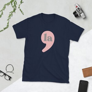 Puede incluir: Una camiseta azul marino con una coma rosa y la palabra "la" en gris.