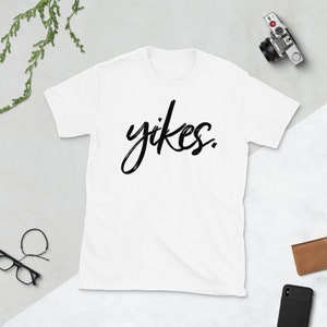 Może przedstawiać: Biała koszulka z napisem "yikes" napisanym czarnym, ozdobnym pismem.