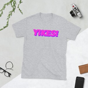 Może przedstawiać: Szary T-shirt z nadrukiem "YIKES!" w różowych i fioletowych literach.