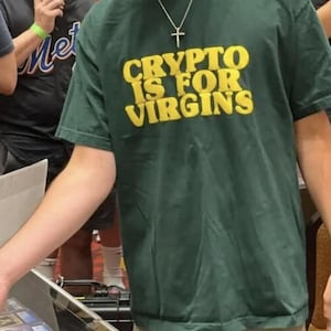 May include: Dark green t-shirt with the text "CRYPTO IS FOR VIRGINS" in yellow block letters. The shirt is being worn by a person. A silver cross necklace is visible.