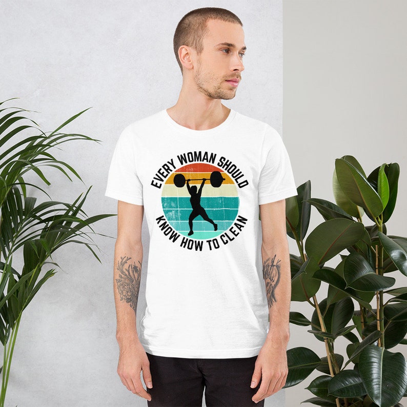 May include: White t-shirt with a retro-style graphic of a woman lifting a barbell. The graphic says "Every Woman Should Know How To Clean".