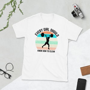 Może przedstawiać: Biała koszulka z grafiką w stylu retro przedstawiającą osobę podnoszącą sztangę z tekstem "Every Girl Should Know How To Clean".