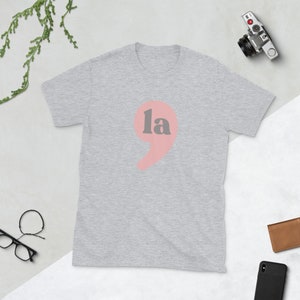 Puede incluir: Una camiseta gris con un punto y coma rosa y la palabra "la" en gris.