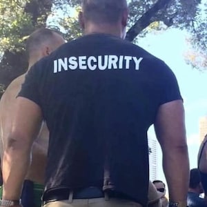 Puede incluir: Una camiseta negra con la palabra "INSECURITY" impresa en letras blancas grandes en la espalda. La camiseta la lleva una persona de pie al aire libre, con otras personas visibles en el fondo.