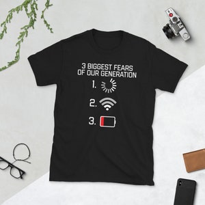 Peut inclure: T-shirt noir avec du texte blanc qui dit "3 Biggest Fears of Our Generation" avec un symbole de chargement, un symbole wifi et un symbole de batterie faible listés comme les trois plus grandes peurs.