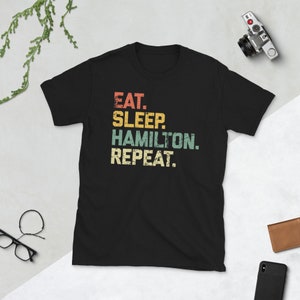 May include: Black t-shirt with a vintage-style graphic that reads "Eat. Sleep. Hamilton. Repeat."