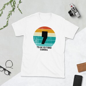 May include: White t-shirt with a retro design. A black comma is set within a circular pattern of orange, yellow, teal, and blue stripes. The text "TEAM OXFORD COMMA" is printed below the circle.
