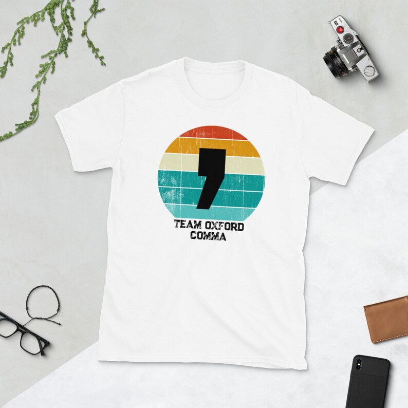 May include: White t-shirt with a retro design. A black comma is set within a circular pattern of orange, yellow, teal, and blue stripes. The text "TEAM OXFORD COMMA" is printed below the circle.