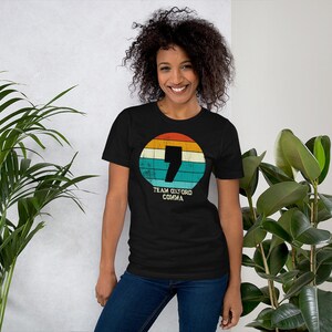May include: Black t-shirt with a retro design. It features a comma inside a circle of orange, yellow, teal, and white stripes. The text "TEAM OXFORD COMMA" is below the circle. The shirt is worn by a person.