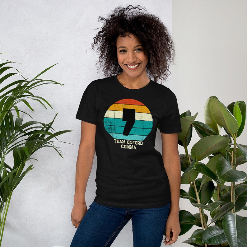 May include: Black t-shirt with a retro design. It features a comma inside a circle of orange, yellow, teal, and white stripes. The text "TEAM OXFORD COMMA" is below the circle. The shirt is worn by a person.