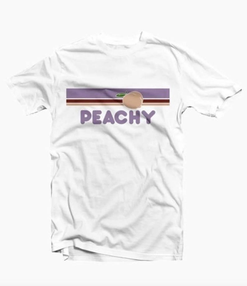 May include: White t-shirt with a graphic design. The design features the word "PEACHY" in purple letters, a peach illustration, and a horizontal stripe design in brown and purple.