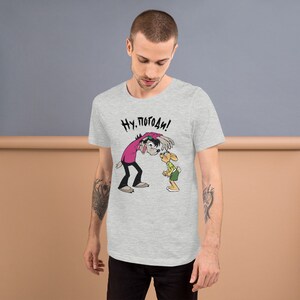 May include: A heather grey t-shirt with a cartoon image of a wolf and a rabbit. The wolf is wearing a pink shirt and the rabbit is wearing a green shirt. The text "Ну. Погоди!" is printed on the shirt.