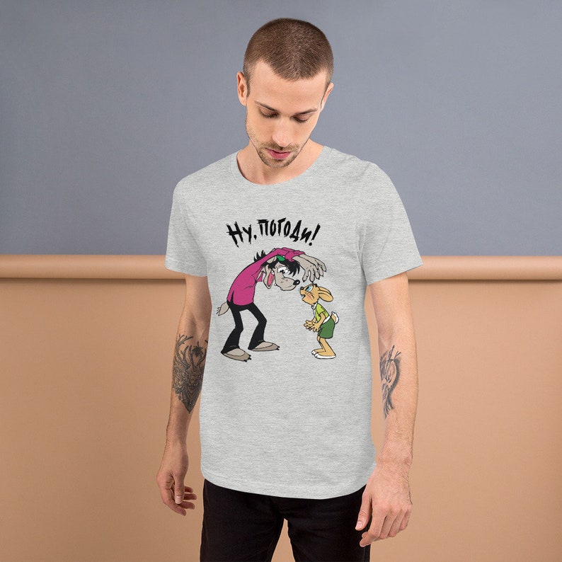 May include: A heather grey t-shirt with a cartoon image of a wolf and a rabbit. The wolf is wearing a pink shirt and the rabbit is wearing a green shirt. The text "Ну. Погоди!" is printed on the shirt.