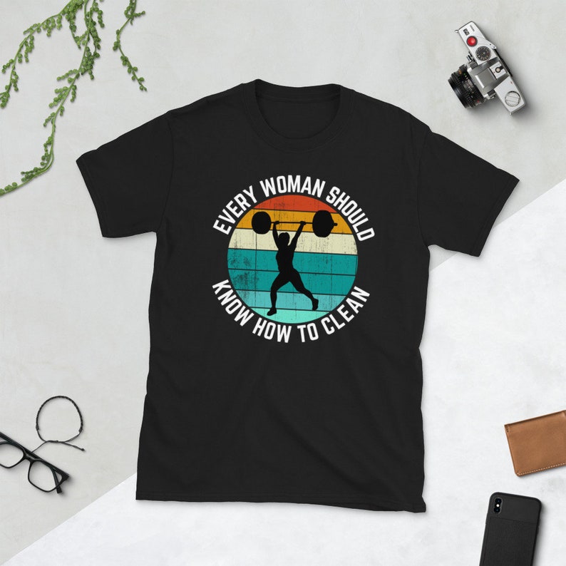 May include: Black t-shirt with a retro-style graphic of a woman lifting a barbell. The graphic is surrounded by the text "Every Woman Should Know How To Clean".