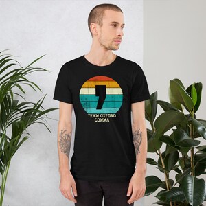 May include: Black t-shirt with a retro design. A comma is inside a circle with orange, yellow, and teal stripes. The text "TEAM OXFORD COMMA" is below the circle. The shirt is made of a soft material.