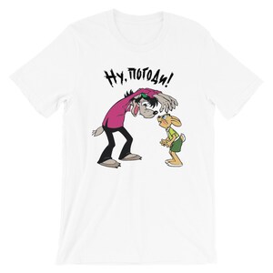May include: White t-shirt with a cartoon image of a wolf and a rabbit. The wolf is wearing a pink shirt and sunglasses and is saying "Ну, погоди!" in Russian.
