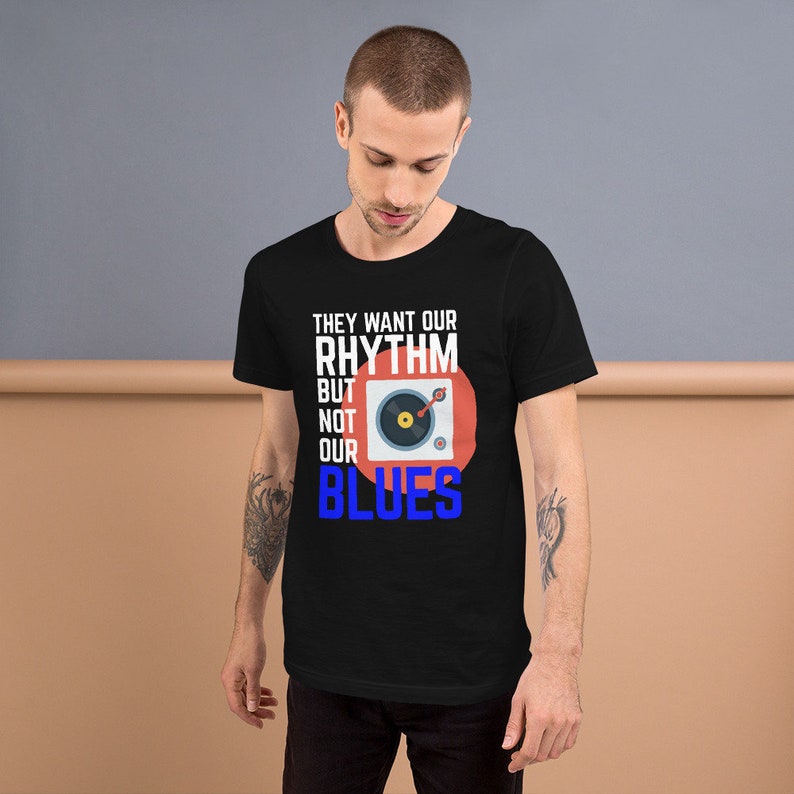 May include: Black t-shirt with a white graphic design that says "They want our rhythm but not our blues". The graphic also includes a red circle with a stylized image of a record player.