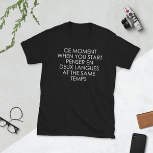 May include: Black t-shirt with white text that reads "CE MOMENT WHEN YOU START PENSER EN DEUX LANGUES AT THE SAME TEMPS".