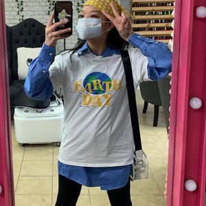 May include: A white t-shirt with the words "Earth Day" in yellow and a blue and green globe design. The shirt is worn over a blue button-down shirt. The person is wearing a yellow patterned headscarf and a face mask.