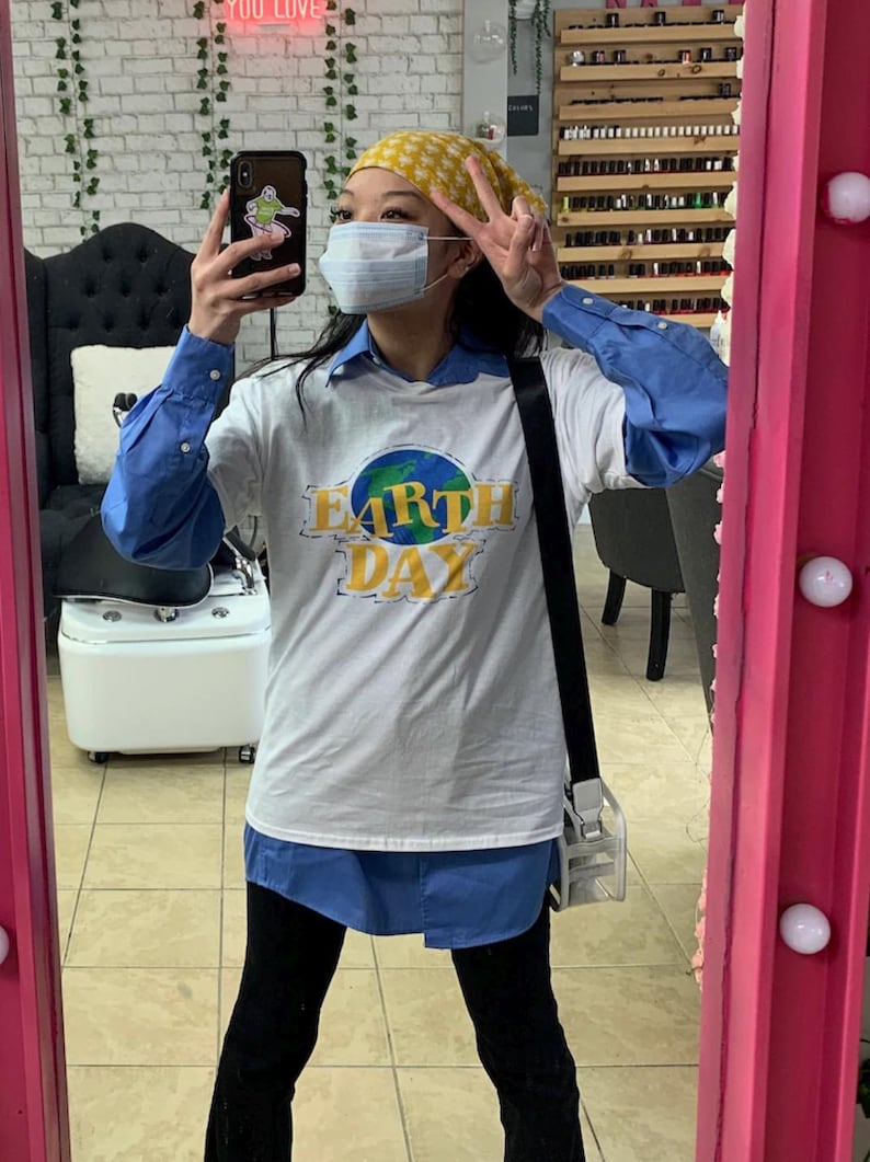 May include: A white t-shirt with the words "Earth Day" in yellow and a blue and green globe design. The shirt is worn over a blue button-down shirt. The person is wearing a yellow patterned headscarf and a face mask.