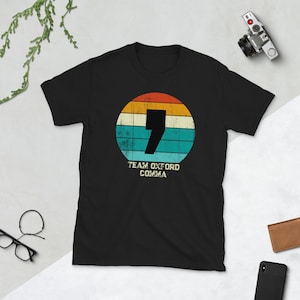 Pode incluir: Camiseta preta com um design retrô. O design inclui um grande número 7 e as palavras "Team Oxford Comma" em branco, dentro de um círculo com listras laranja, amarelo, verde-azulado e azul. Uma câmera, óculos e uma carteira estão no fundo.