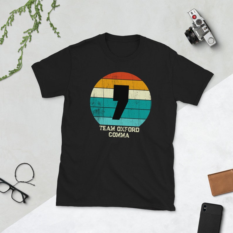 May include: Black t-shirt with a retro design. The design includes a large number 7 and the words "Team Oxford Comma" in white, set within a circle with orange, yellow, teal, and blue stripes. A camera, glasses, and a wallet are in the background.