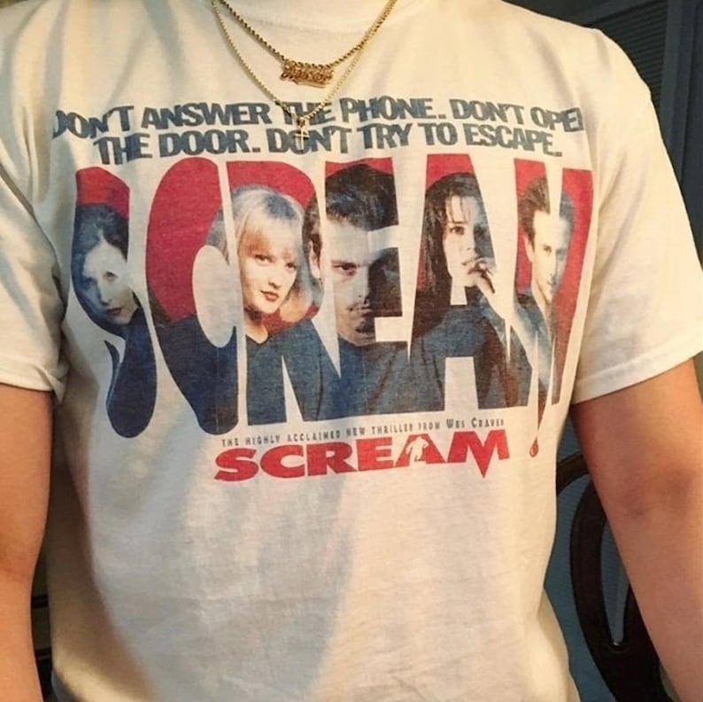 May include: Cream-colored t-shirt featuring the word "SCREAM" in red and blue, with images of characters from the movie. The shirt also has the text "DON'T ANSWER THE PHONE. DON'T OPEN THE DOOR. DON'T TRY TO ESCAPE."