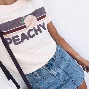 May include: White t-shirt with a peach graphic and the word "PEACHY" in a bold font. The shirt has a pink neckline and short sleeves. Paired with a blue denim skirt.