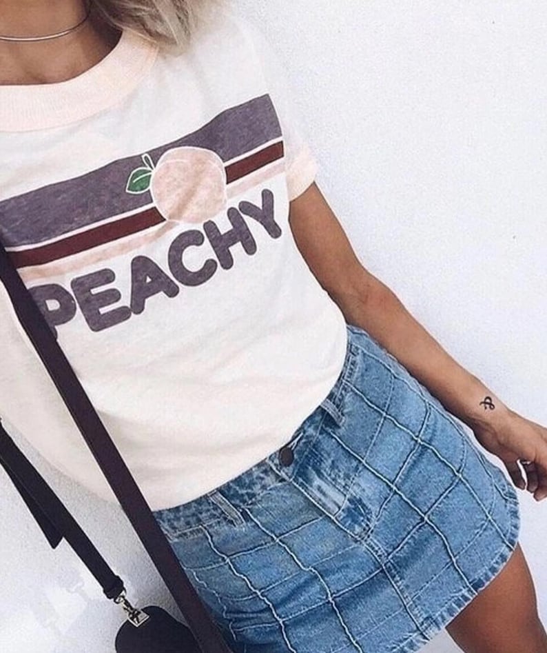 May include: White t-shirt with a peach graphic and the word "PEACHY" in a bold font. The shirt has a pink neckline and short sleeves. Paired with a blue denim skirt.