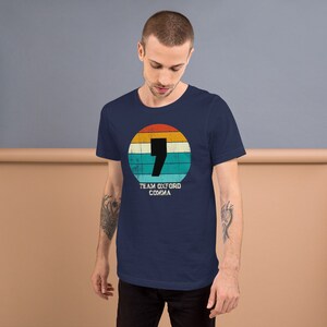 May include: A navy blue t-shirt with a retro design. The design features a comma inside a circle with orange, yellow, and blue stripes. The text "TEAM OXFORD COMMA" is below. The shirt is made of soft material.