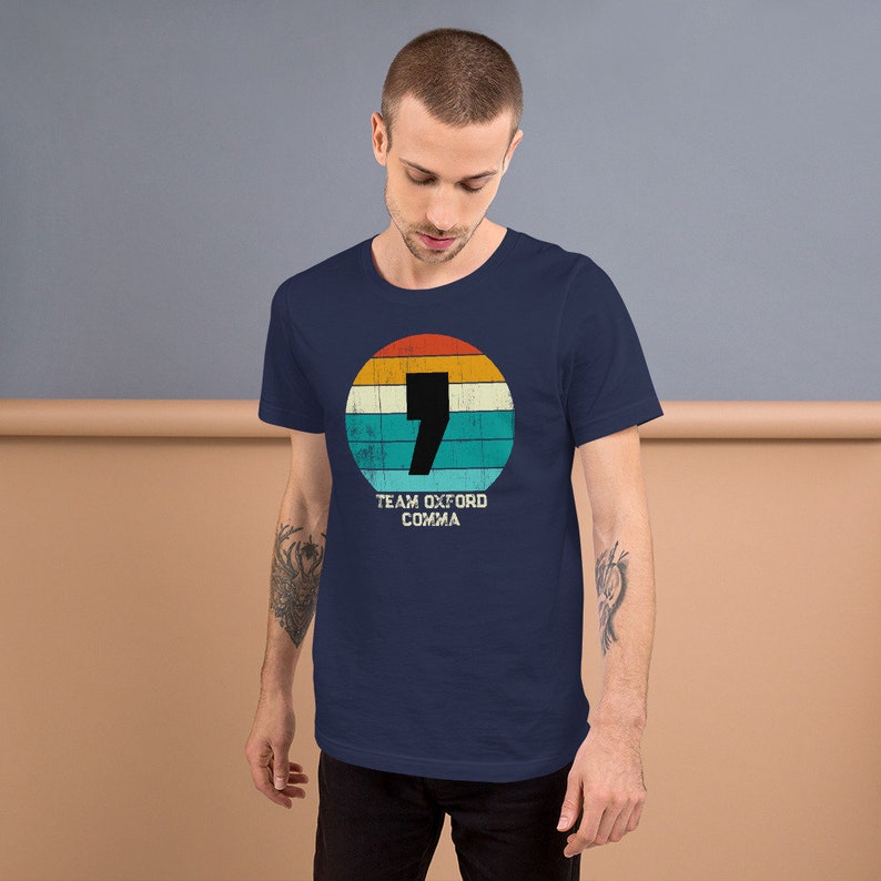 May include: A navy blue t-shirt with a retro design. The design features a comma inside a circle with orange, yellow, and blue stripes. The text "TEAM OXFORD COMMA" is below. The shirt is made of soft material.