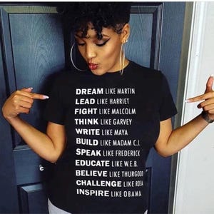 May include: Black t-shirt with white text listing inspirational figures and their qualities: Dream like Martin, Lead like Harriet, Fight like Malcolm, Think like Garvey, Write like Maya, Build like Madam C.J., Speak like Frederick, Educate like W.E.B., Believe like Thurgood, Challenge like Rosa, Inspire like Obama.
