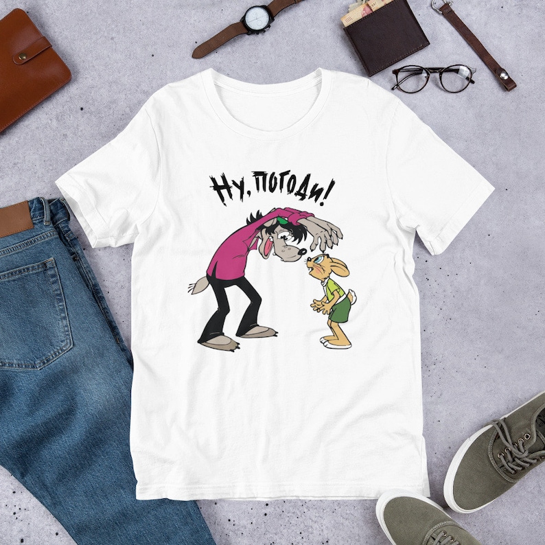 May include: White t-shirt with a cartoon image of a wolf and a rabbit. The wolf is wearing sunglasses and a pink shirt. The rabbit is wearing a green shirt and green shorts. The text "Ну, погоди!" is printed on the shirt.