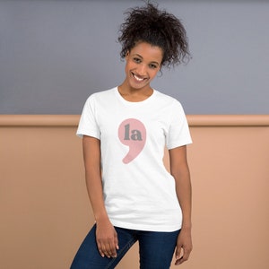 Puede incluir: Una camiseta blanca con un punto y coma rosa y la palabra "la" en gris.