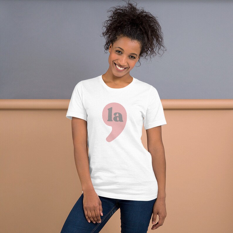 Puede incluir: Una camiseta blanca con un punto y coma rosa y la palabra "la" en gris.