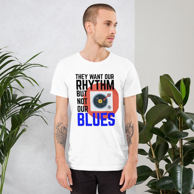 May include: White t-shirt with black text that reads "They want our rhythm but not our blues". A red circle with a black record player inside is next to the text.