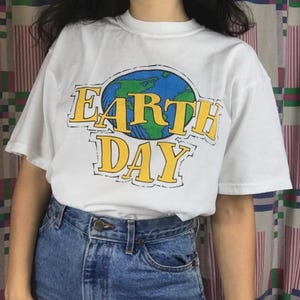 May include: White t-shirt with a crew neck and short sleeves. The shirt features a graphic of the Earth in blue and green, with the words "EARTH DAY" in yellow block letters.