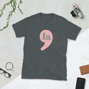 Puede incluir: Una camiseta gris oscuro jaspeada con un signo de puntuaci&oacute;n rosa y la palabra "la" en gris.