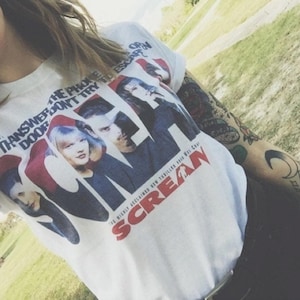 May include: White t-shirt featuring a movie poster design for the film "Scream." The graphic includes images of the cast and the film's title in red. The shirt also has the text "The Phone Don't Try To Escape" printed on it.