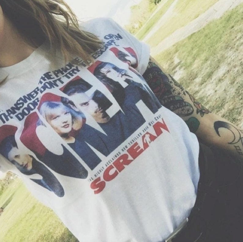 May include: White t-shirt featuring a movie poster design for the film "Scream." The graphic includes images of the cast and the film's title in red. The shirt also has the text "The Phone Don't Try To Escape" printed on it.