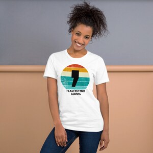May include: White t-shirt with a retro design. It features a black comma inside a circular pattern of orange, yellow, and blue stripes. The text "TEAM OXFORD COMMA" is printed below. The t-shirt is a crew neck style.