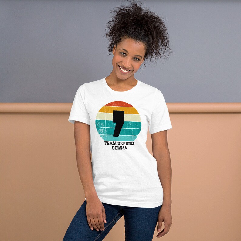 May include: White t-shirt with a retro design. It features a black comma inside a circular pattern of orange, yellow, and blue stripes. The text "TEAM OXFORD COMMA" is printed below. The t-shirt is a crew neck style.