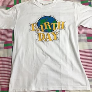 May include: White t-shirt featuring a graphic for Earth Day. The design includes a blue and green globe with the words "EARTH DAY" in yellow, outlined in black. The shirt is made of a soft material and has a crew neck.